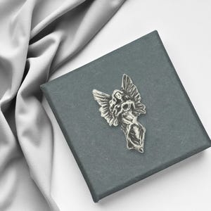 May include: A silver angel pin with outstretched wings rests on a dark grey box. The pin features a detailed angel figure. A grey fabric is draped in the background.