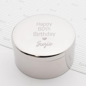 Könnte beinhalten: Eine silberfarbene, runde Schmuckschatulle mit Deckel. Der Deckel ist mit "Happy 60th Birthday Suzie" und einem Herzsymbol graviert. Die Schatulle steht auf einer weißen Oberfläche mit einem sich wiederholenden Muster aus Herzen und dem Wort "love".