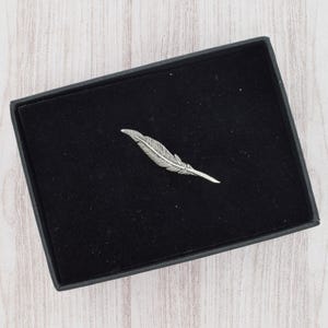 May include: A silver feather-shaped brooch displayed in an open black box with a black interior. The brooch has detailed lines to represent the feather's structure. The box is open, showing the brooch against the dark background.