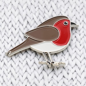 May include: A metal enamel pin shaped like a robin bird. The bird features a bright red chest, brown wings and back, and a white belly. It has a small black eye and a pointed black beak. The pin is outlined in silver metal and has two small feet.