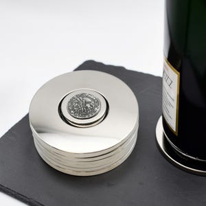 Op de afbeelding: Een stapel ronde onderzetters van gepolijst zilverkleurig metaal met een centraal medaillon met een reliëf van een historische figuur te paard. De onderzetters hebben een glad, reflecterend oppervlak naast een donkere glazen fles.