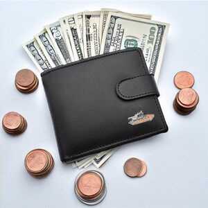 May include: A black leather wallet with a small tank design on the front. The wallet is open and filled with US dollar bills. There are stacks of copper coins scattered around the wallet.