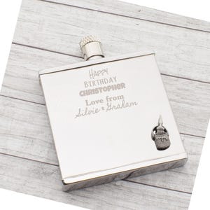May include: A silver hip flask with a screw-on cap. The front of the flask is engraved with the words "Happy Birthday Christopher, Love from Silvie & Graham". A small decorative paint brush charm is attached to the lower right corner.