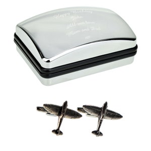 Puede incluir: Una caja rectangular plateada con tapa curvada y borde negro. La tapa tiene texto grabado. Debajo, dos gemelos plateados con forma de avión.