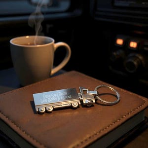 Peut inclure: Un porte-clés argenté en forme de camion avec le texte gravé "DRIVE SAFE DADDY LOVE BOBBY & KELLY XX" repose sur une surface en cuir marron. Une tasse de café est à proximité, avec de la vapeur qui s'élève.