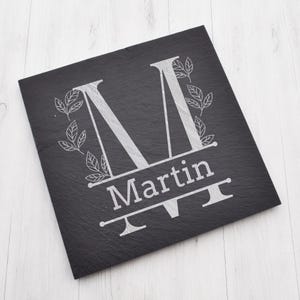Puede incluir: Un letrero de pizarra negra con una letra M grabada en plata y el nombre Martin. La letra M está rodeada de un diseño floral.