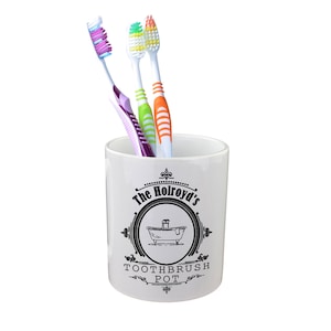 Puede incluir: Portacepillos de dientes de cerámica blanca con texto negro que dice "The Holroyd's Toothbrush Pot" y una ilustración en blanco y negro de una bañera. Tres cepillos de dientes están en el portacepillos, uno morado, uno verde y uno naranja.