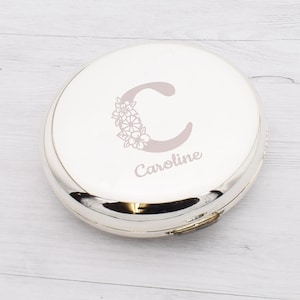 Op de afbeelding: Een zilveren compacte spiegel met een witte bovenkant. De bovenkant heeft een bloemenontwerp rond de letter "C" en de naam "Caroline" in lichtgrijs. De compact heeft een glanzend, reflecterend oppervlak.