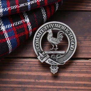 May include: A silver-colored circular emblem with a rooster in the center, the words "COMMIT THY WORK TO GOD" are inscribed around the edge. The emblem rests on a wooden surface next to a folded plaid fabric.