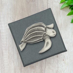 May include: A pewter-coloured turtle figurine with detailed shell lines and flipper-like limbs rests on a dark grey box. The turtle is a decorative item, possibly a paperweight or small sculpture. The background is a light wood surface.