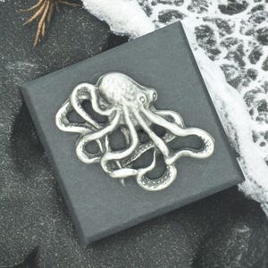 May include: A silver octopus brooch with detailed tentacles and a textured finish. The brooch is presented on a dark grey box, possibly for jewellery or accessories. The design has a nautical theme.