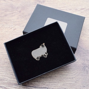 Pode incluir: Um broche de esmalte preto e branco de uma ovelha em uma caixa preta com interior de veludo preto.
