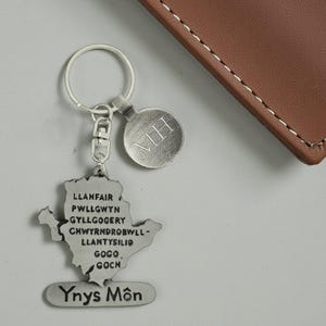 May include: A silver-colored keychain with a map-shaped pendant featuring text and a round tag with "MH" engraved. The keychain is attached to a key ring and a clasp. The text on the pendant reads "LLANFAIR PWLLGWYN GYLLGOGERY CHWYRNDROBWLL-LLANTYSILIO GOGO, GOCH YNYS Môn."