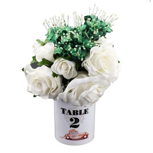 以下が含まれることがあります： 白いバラと緑の葉を、白い陶器の花瓶に入れています。花瓶には「TABLE 2」の文字とピンクの車の絵が描かれています。