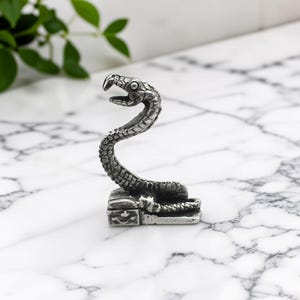 May include: A silver-toned metal figurine of a snake with a textured, scaled body and an open mouth, coiled atop a small rectangular base that features a subtle embossed crown detail on its front side, finished with a dark, weathered patina.