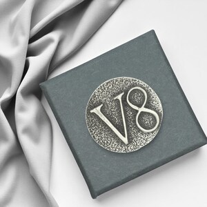 Op de afbeelding: Een zilverkleurig metalen embleem met de letters "V8" op een textuurrijke, cirkelvormige achtergrond. Het embleem is geplaatst op een donkergrijze vierkante doos, met grijze stof op de achtergrond.