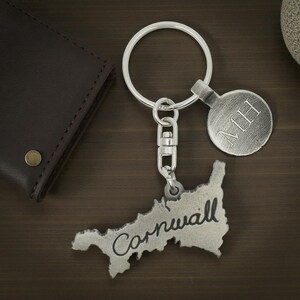 May include: A silver-colored keychain featuring the outline of Cornwall, with the word "Cornwall" in a handwritten font. The keychain includes a round tag with the initials "MH". A dark brown leather wallet is visible in the background.