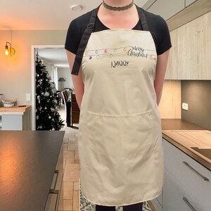 May include: A white apron with a black strap and a pocket. The apron has a design of colorful Christmas lights and the text "Merry Christmas Nanny".