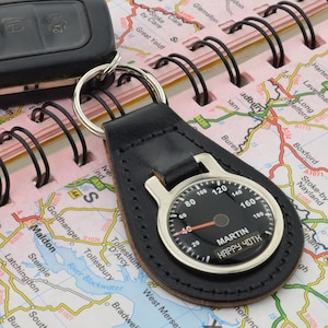 May include: Black leather keychain with a silver speedometer-style charm. The charm has a black face with white numbers and a red needle. The text "MARTIN HAPPY 40TH" is printed on the face.