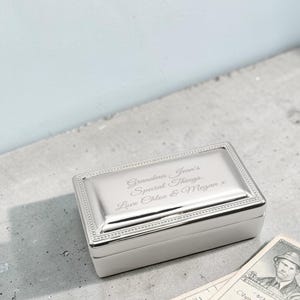 Puede incluir: Una caja rectangular plateada con tapa abisagrada. La tapa está grabada con las palabras "Grandma Jean's Special Things. Love Chloe & Megan x". La caja tiene un borde de cuentas y está sobre una superficie gris.