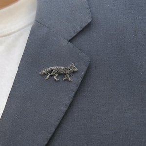 May include: A metallic silver-toned fox lapel pin featuring a detailed, textured fur design. The fox is depicted in a walking pose with its tail extended behind it. The pin is attached to the dark grey fabric of a structured blazer lapel.