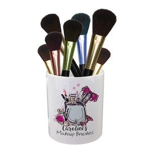 May include: White ceramic makeup brush holder with a colourful illustration of makeup supplies and the text "Caroline's Makeup Brushes". The holder is filled with a variety of makeup brushes.