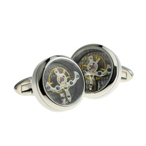 May include: A pair of silver cufflinks with a clear circular face revealing a working watch mechanism. The watch face has a black and white carbon fiber pattern with gold gears and blue accents.