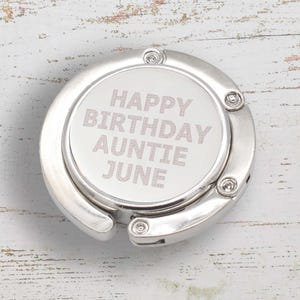 以下が含まれることがあります： 白い背景にピンクの文字で「Happy Birthday Auntie June」と書かれた銀色の金属製のバッグチャームです。