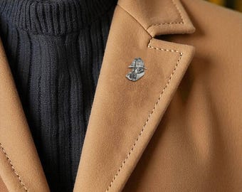 May include: A silver-colored lapel pin depicting a person wearing a safari hat and holding a globe. The pin is attached to a tan blazer, with a dark blue sweater visible underneath. The blazer has a classic notched lapel and a stitched edge.