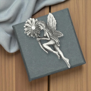 May include: A silver fairy brooch holding a flower with a central crystal. The fairy has detailed wings and flowing hair. The brooch is set on a dark grey box, with a light blue fabric and wooden surface in the background.