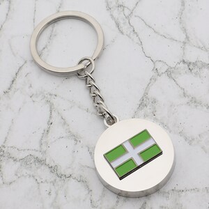 May include: A silver keychain with a round charm featuring the green and white flag of Cornwall. The charm is attached to a silver chain and a silver key ring.