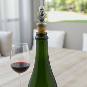 Può includere: Una bottiglia di vino verde con un tappo a forma di busto color argento. Un bicchiere di vino rosso è a sinistra. Il tappo presenta una scultura dettagliata della testa e delle spalle di un uomo. La bottiglia è su un tavolo.