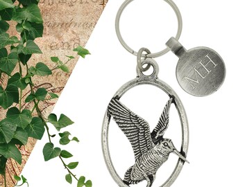 Puede incluir: Llavero plateado con un diseño de pájaro detallado en un marco ovalado. Una etiqueta redonda con las iniciales "MH" está unida al llavero.