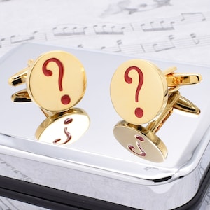 Puede incluir: Un par de gemelos de oro con un signo de interrogación rojo. Los gemelos están sobre una superficie plateada.