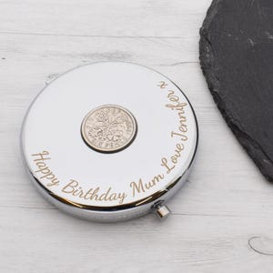 Puede incluir: Un espejo compacto plateado con una moneda de seis peniques británica de 1953 incrustada en el centro. El espejo está grabado con el texto "Happy Birthday Mum Love Jennifer x".
