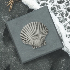 May include: A silver-colored seashell-shaped pendant or brooch rests on a dark gray square box. The seashell has detailed lines radiating from the center. The background includes a textured surface with white and black patterns.