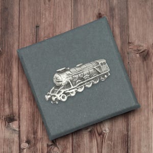 Pode incluir: Um alfinete de trem prateado repousa sobre uma caixa cinza escuro, contra um fundo de madeira. O alfinete retrata uma locomotiva a vapor detalhada com rodas e chaminé. A caixa fornece um fundo contrastante.