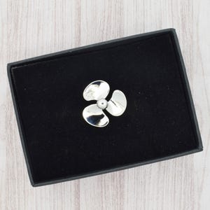 Peut inclure: Broche argentée en forme d'hélice à trois pales incurvées et un centre perlé. La broche est présentée dans un écrin noir avec un intérieur en velours, sur un fond clair en bois.