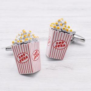 Könnte beinhalten: Ein Paar silberne Manschettenknöpfe mit einem Popcorn-Box-Design. Die Boxen sind weiß mit roten Streifen und den Worten "Pop Corn" in rot.