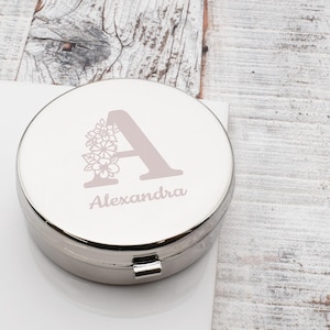 May include: A silver-colored, round pill box with a hinged lid. The lid features a large letter "A" in a light mauve color, adorned with floral accents. The name "Alexandra" is printed below the letter in a matching font.
