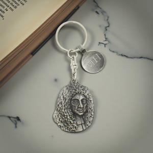 May include: A silver-coloured keyring featuring a portrait of a man with long, curly hair. The keyring includes a round tag with the initials "MH" engraved. The background is a marble-like surface.