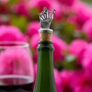 May include: A decorative bottle stopper featuring a silver-coloured bagpipe player design. The stopper is on a green glass bottle with a cork. The background is a blurred view of pink flowers and a wine glass.