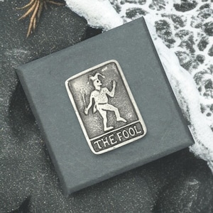 May include: A silver-colored metal pin featuring a stylized illustration of a jester holding a staff, with the text "THE FOOL" at the bottom. The pin is set on a dark gray box, with a textured background.