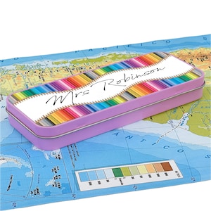 Puede incluir: Un estuche rectangular lila con una tapa que presenta un diseño de lápices de colores. La tapa tiene el texto "Mrs Robinson" en cursiva. El estuche está sobre un mapa.