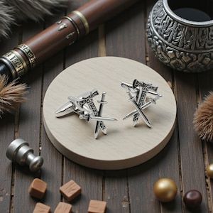 May include: Silver cufflinks with a crossed swords design, displayed on a light wooden surface. The cufflinks have intricate detailing and a polished finish. The background includes a wooden table, a pen, and decorative items.
