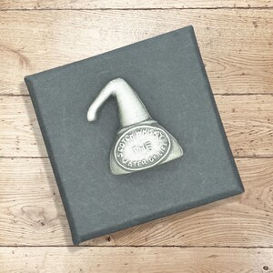 May include: A silver-coloured metal pin in the shape of a whisky still, with the words "Scotch Whisky The Water of Life" inscribed. The pin is on a dark grey box, set on a wooden surface.