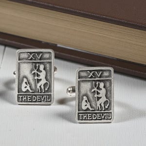 Puede incluir: Un par de gemelos plateados con un diseño rectangular. Cada gemelo presenta la imagen de la carta del tarot del Diablo. El número romano XV está en la parte superior y las palabras "THE DEVIL" en la parte inferior.