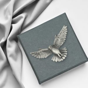 May include: A silver owl brooch with outstretched wings sits on a dark grey box. The brooch is detailed with feathers. A grey fabric is draped in the background. The brooch is a decorative item.