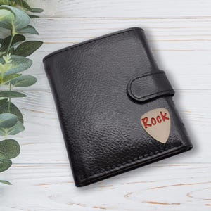 May include: Black leather wallet featuring a guitar pick design. The pick is beige with the word "Rock" in red. The wallet has a snap closure and a textured surface. The background is a white wooden surface with green leaves.