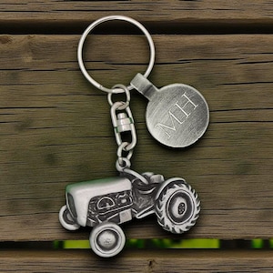 Könnte beinhalten: Ein silberfarbener Traktor-Schlüsselanhänger mit einem runden Anhänger, auf dem die Initialen "MH" eingraviert sind. Der Traktor hat detaillierte Räder und ein klassisches Design. Der Schlüsselanhänger ist an einem silbernen Ring befestigt.